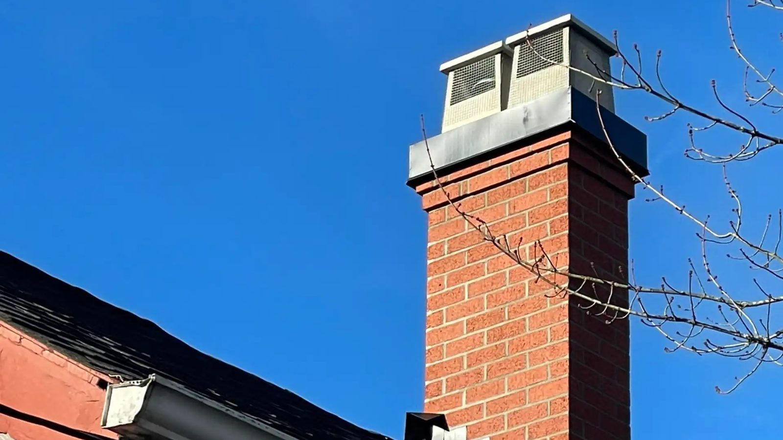 A red-brick chimney with a black metal cap and two screened chimney caps against a clear blue sky, from a finished Lindbjerg Chimney job