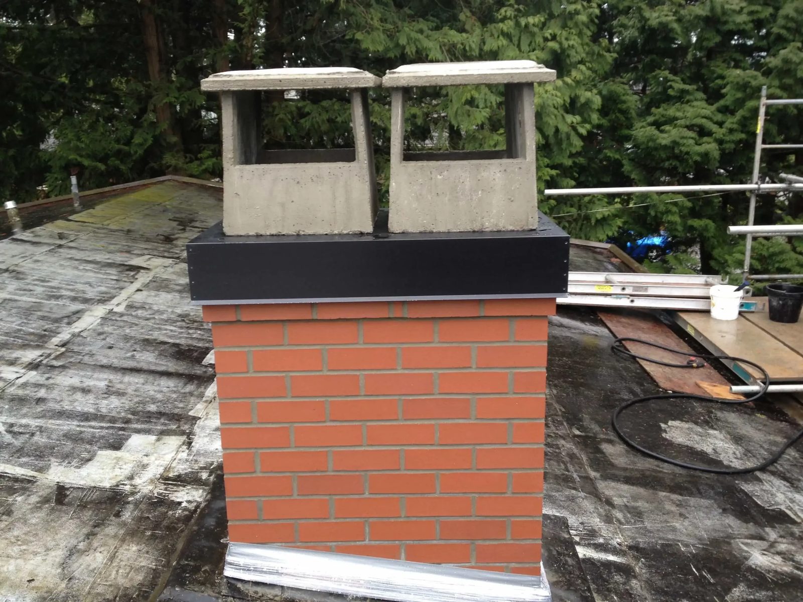 A freshly bricked orange-red chimney with a black metal cap and two concrete chimney pots on a flat roof, with scaffolding and a ladder behind it