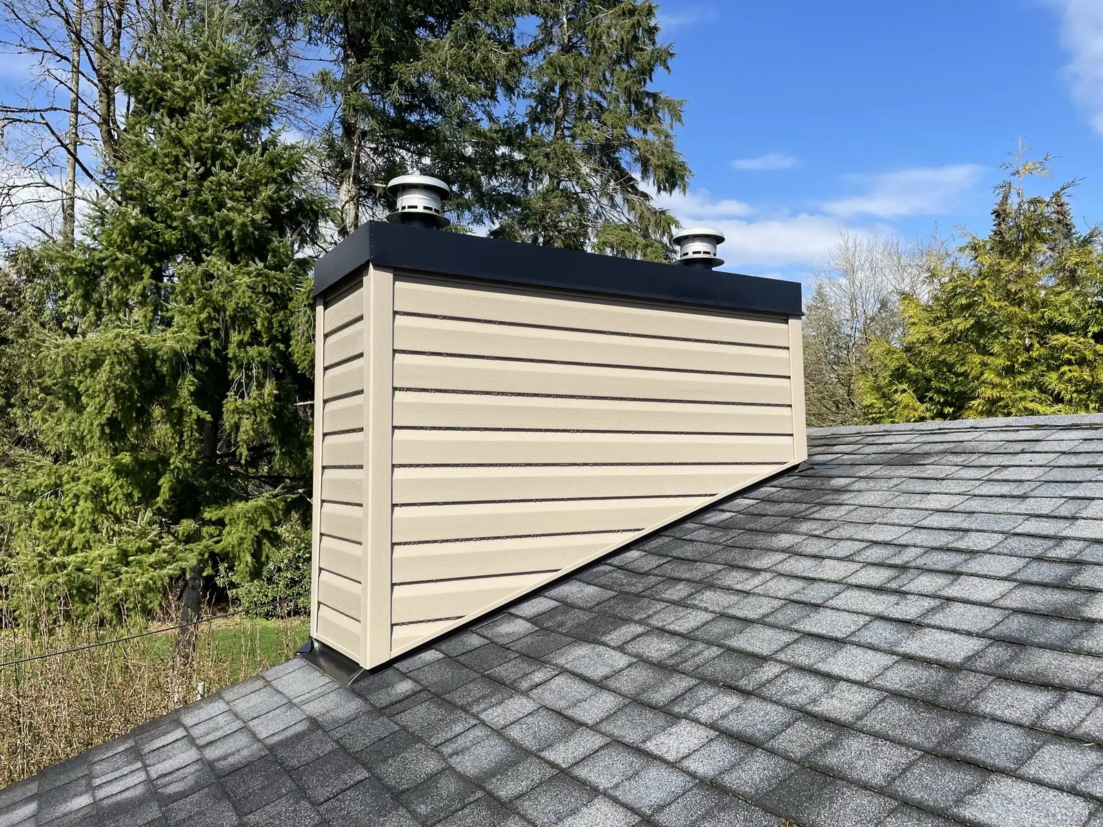 A chimney wrapped in tan horizontal siding with a black cap and two round vent caps, standing on a grey shingle roof with evergreens behind