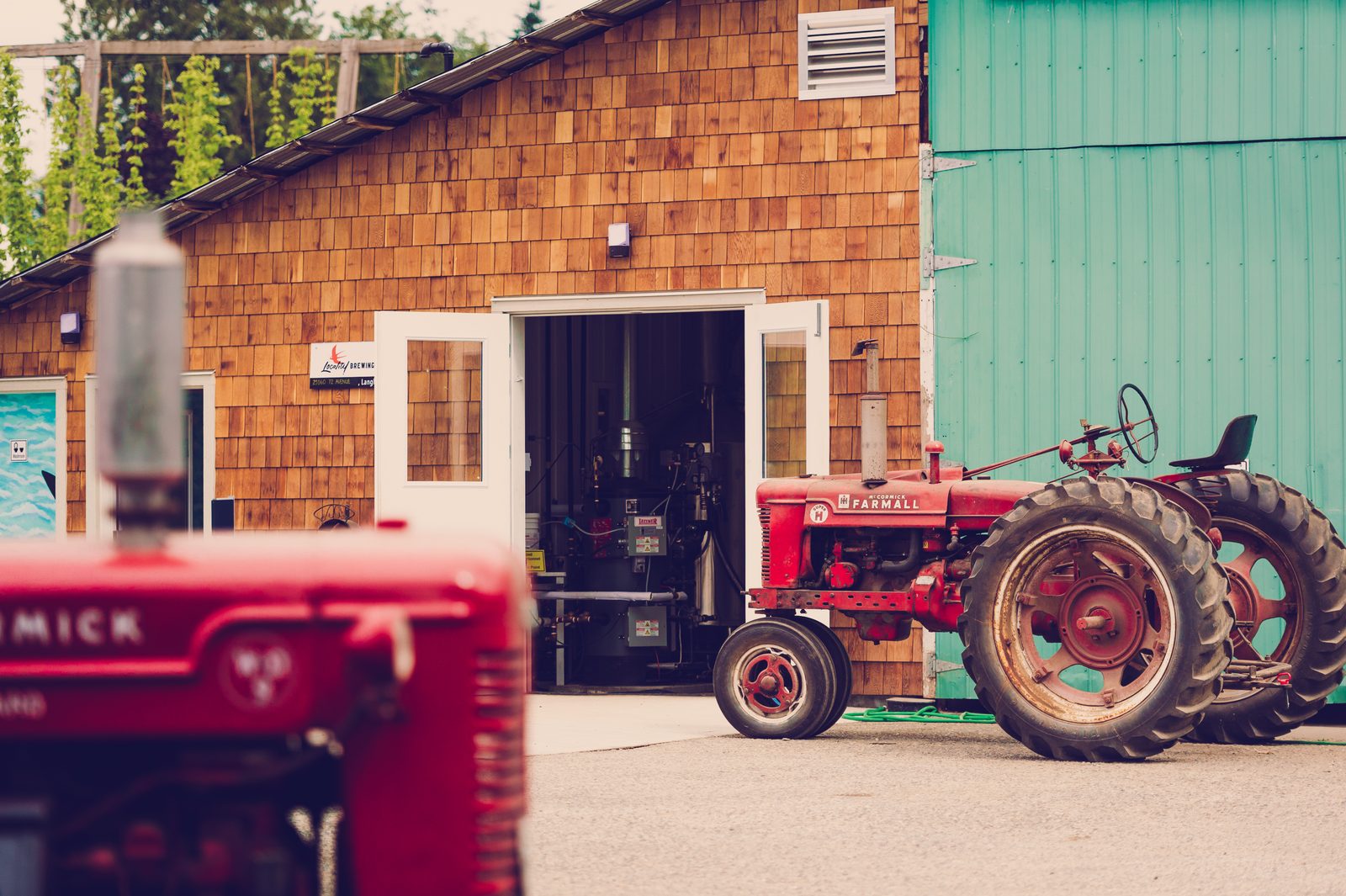 The cedar-shingled brewhouse at Locality Brewing with its doors open onto the brewing tanks, and two red vintage tractors parked in front of a green barn