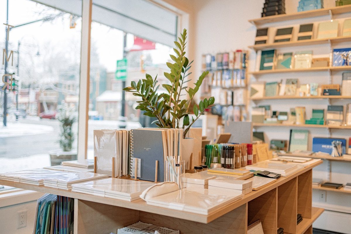 Inside The Mailroom in Fort Langley: wooden display tables of notebooks and pens in front of shelves of paper goods, with the village street through the front window