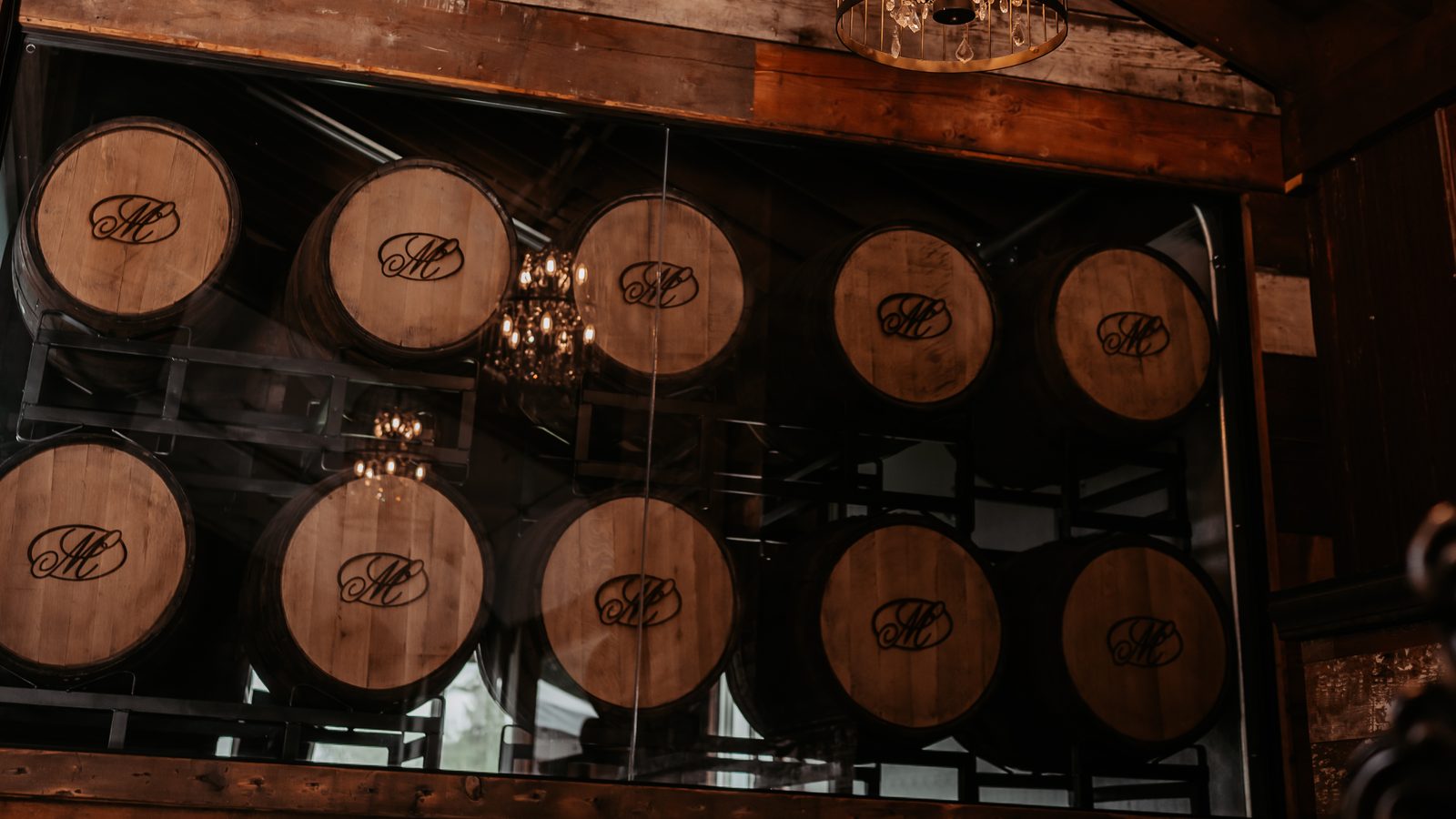 A wall of oak barrels, each branded with a looped M, stacked behind glass under a chandelier at the Manners Cider house