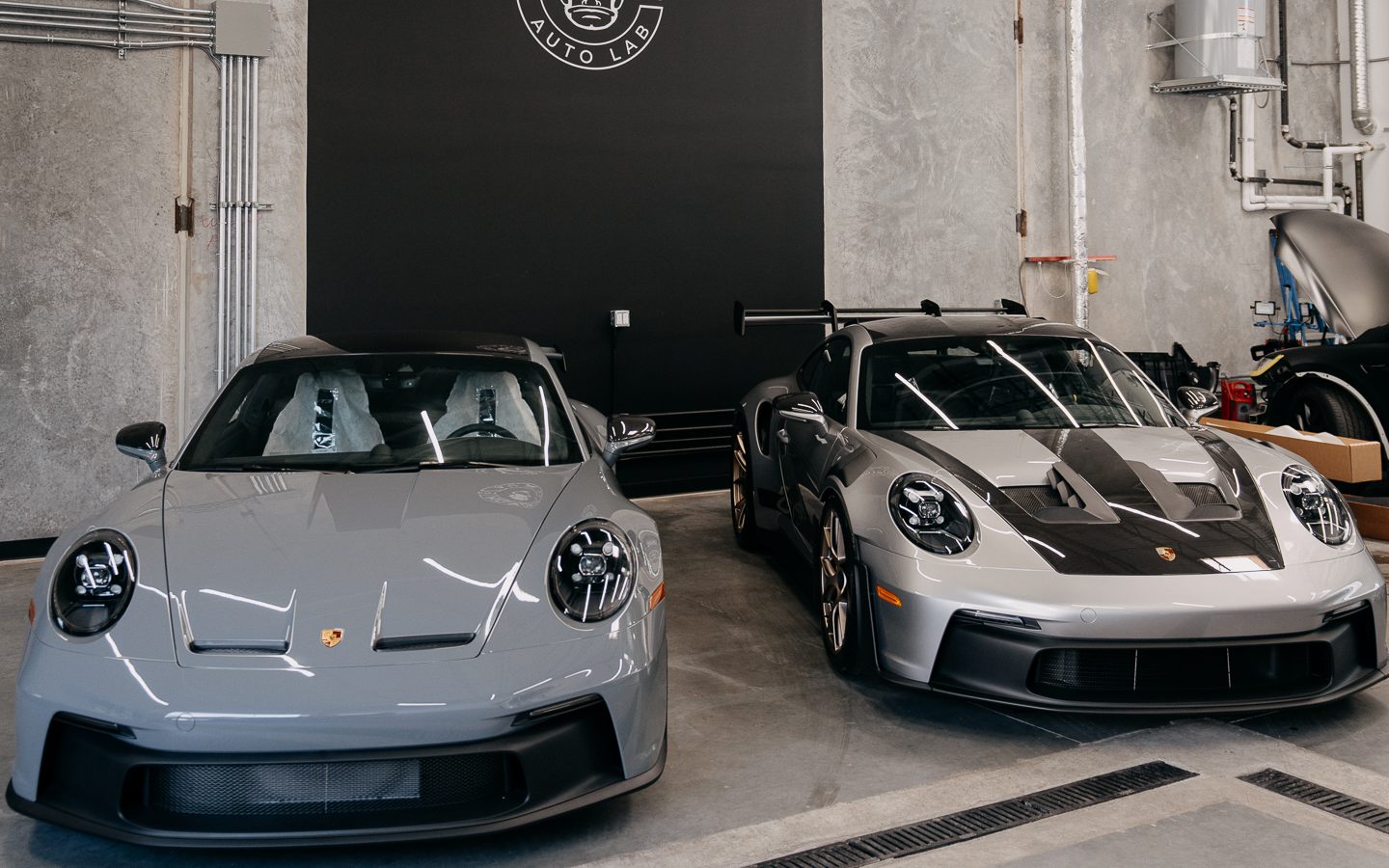 Two Porsche 911 GT3 cars parked inside the Monster Auto Lab workshop in front of a black logo wall on raw concrete, with exposed pipes and ductwork overhead