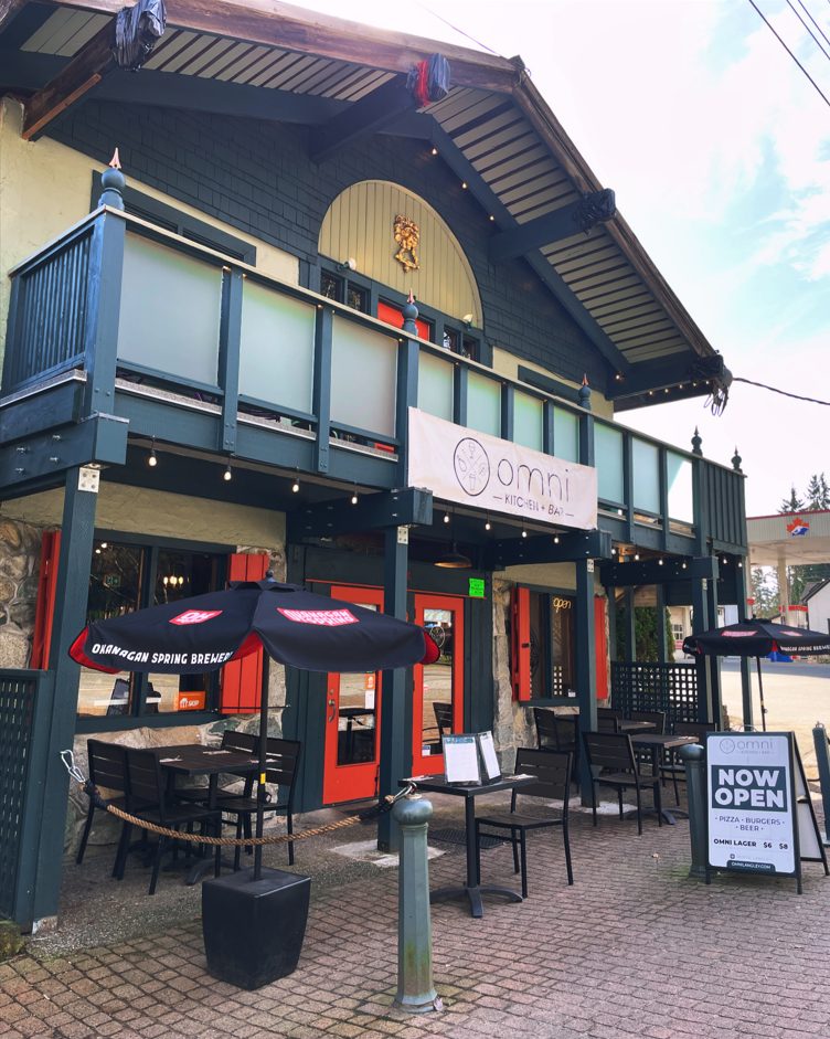 Omni Kitchen + Bar's storefront on Glover Road in Fort Langley: a dark green two-storey building with a balcony, orange doors, patio tables under black umbrellas and a Now Open sandwich board