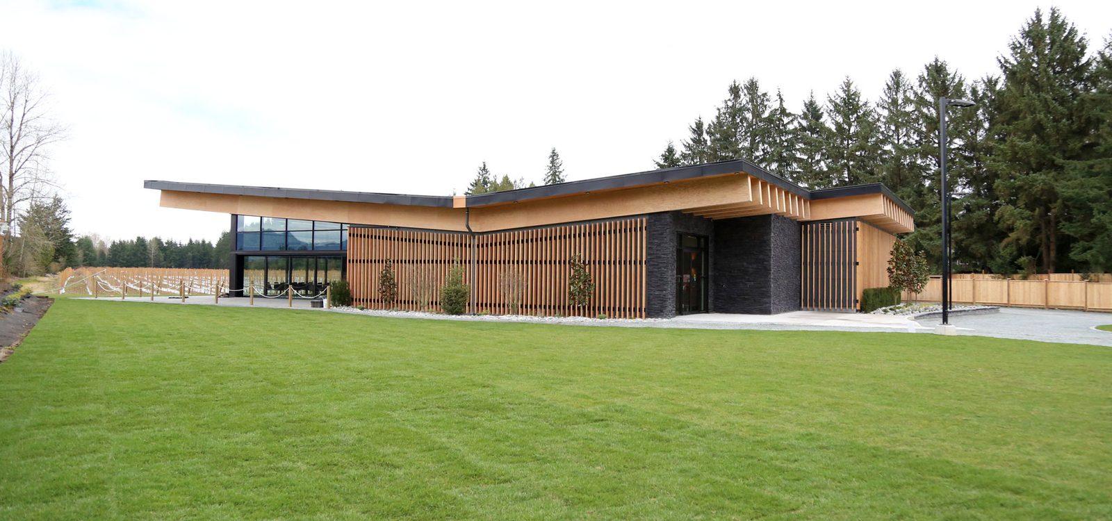 The Otter Trail Winery building on 248 Street: a low timber-and-black-stone building with a sweeping roofline, vertical wood slats, a glass dining room at the left and a mown lawn in the foreground