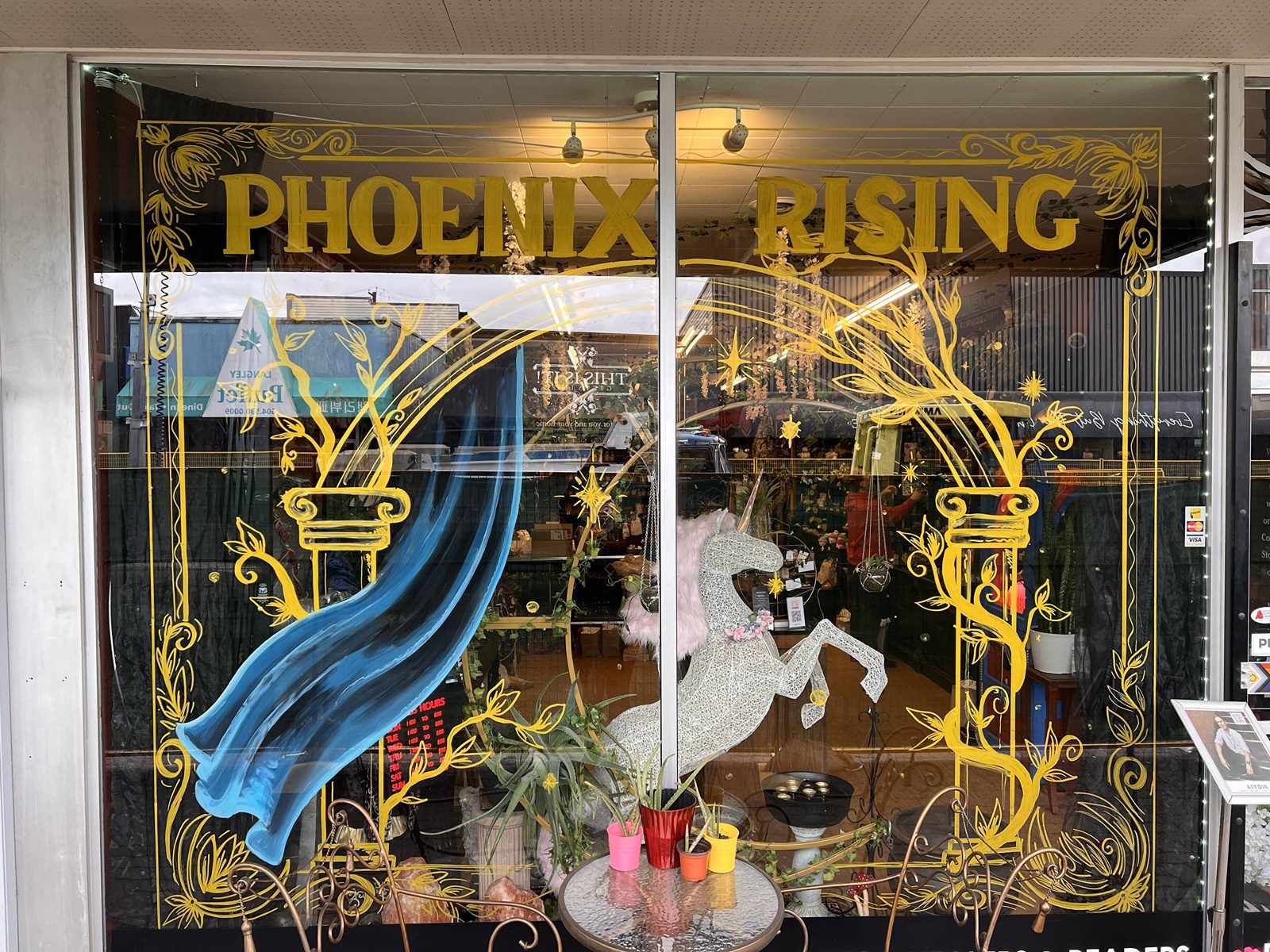 The Phoenix Rising shop window on Fraser Highway, painted with gold lettering and a blue phoenix wing, with a sparkly unicorn figure and plants on display inside
