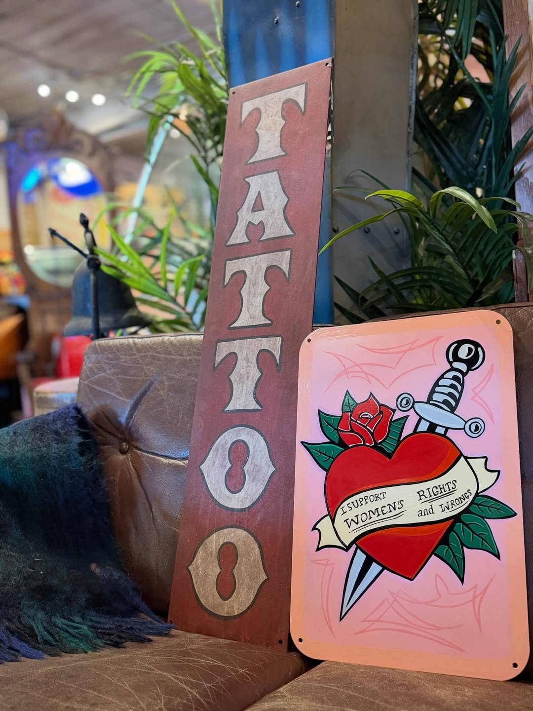 A tall hand-painted TATTOO sign and a heart-and-dagger sign propped up inside Rempel Mercantile, with plants and shop displays behind