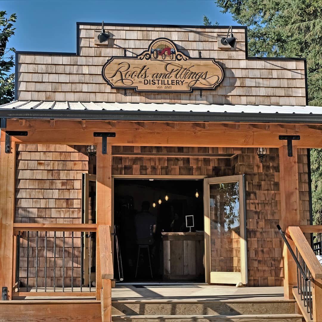 The cedar-shingled Roots and Wings Distillery tasting room, with the carved wooden sign above an open double doorway and wooden railings