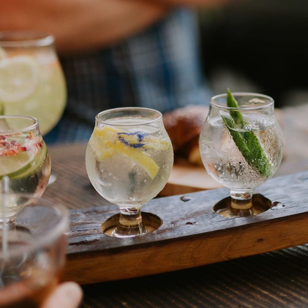 A barrel-wood paddle holding a flight of garnished gin-and-soda tasting glasses, with lemon, lime, raspberry and cucumber