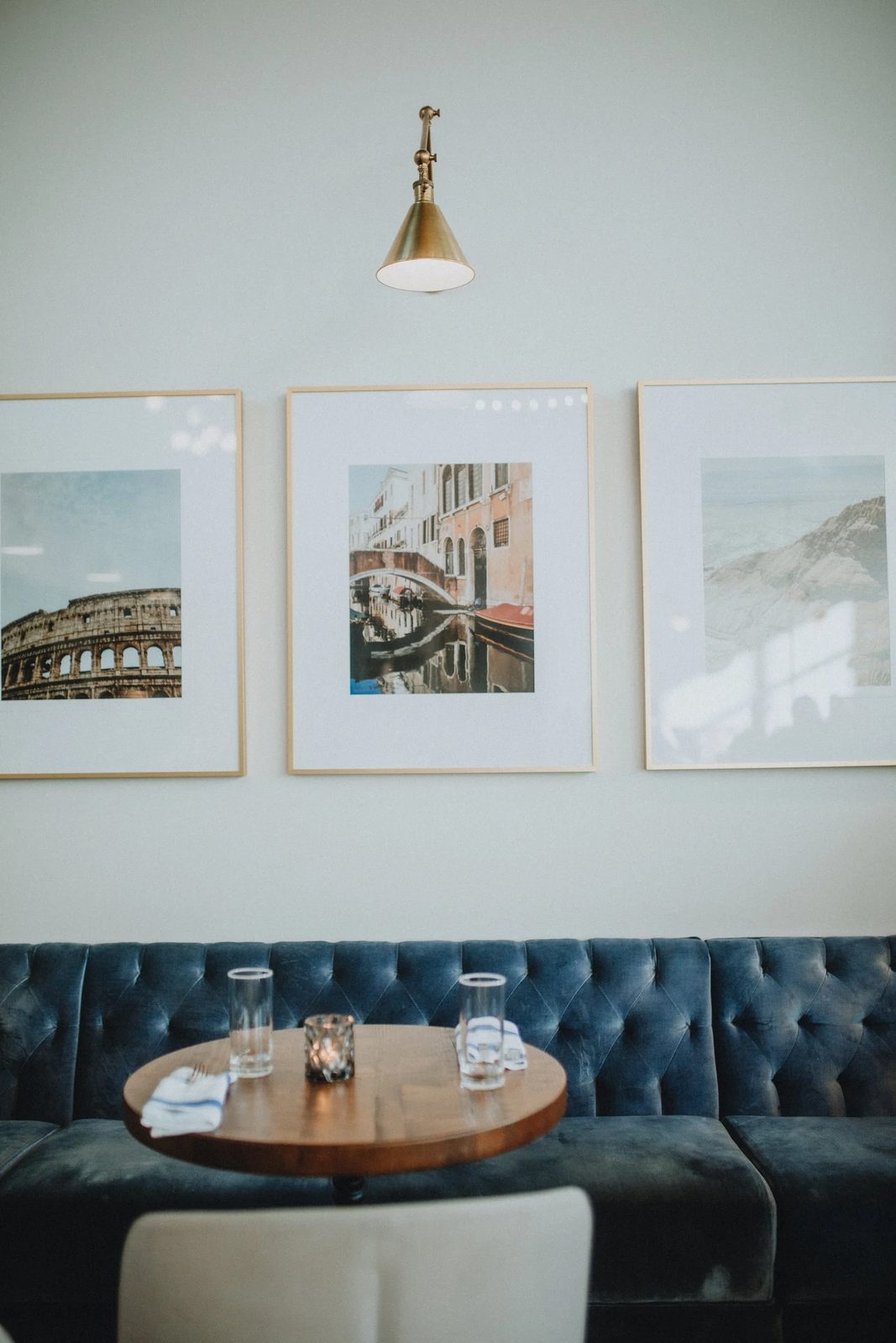 A corner table in Sabà Bistro: a round wooden table with two water glasses and a candle in front of a navy tufted velvet banquette, under three framed prints of Rome, Venice and a coastline and a brass wall lamp