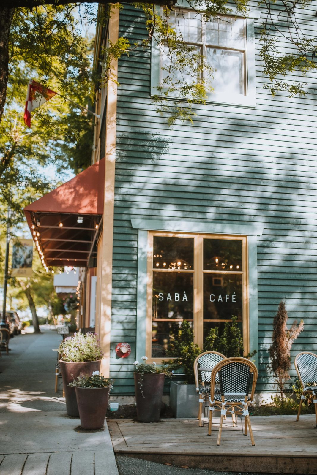 The corner of the Sabà Cafe building in Fort Langley: green clapboard walls, a window lettered SABÀ CAFÉ, a red awning strung with lights, flower pots and bistro chairs on a small wooden deck