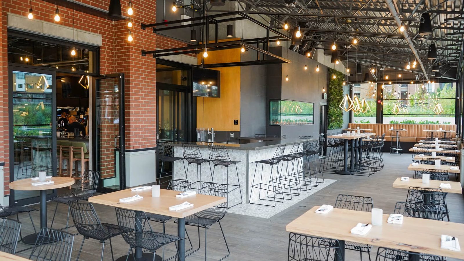 The covered patio at Sidebar Grill with pale wood tables, black wire chairs, strings of bare bulbs, a brick wall, a tiled bar with stools and glass doors opening to the inside