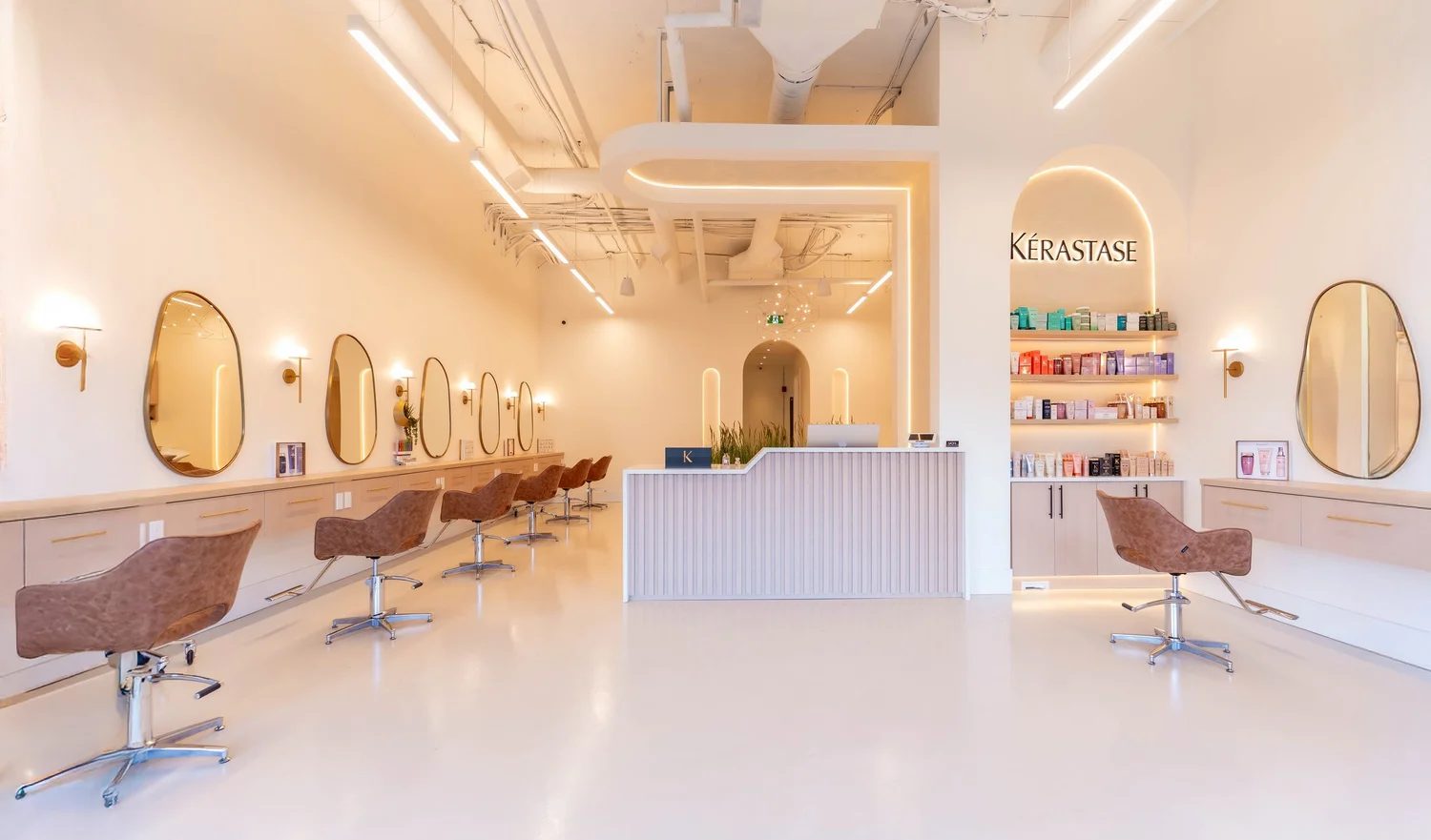 The floor of Skye Hair Salon in Willoughby: a row of tan leather chairs under oval brass-framed mirrors, a pale fluted reception desk and a lit arched alcove of Kérastase haircare at the back
