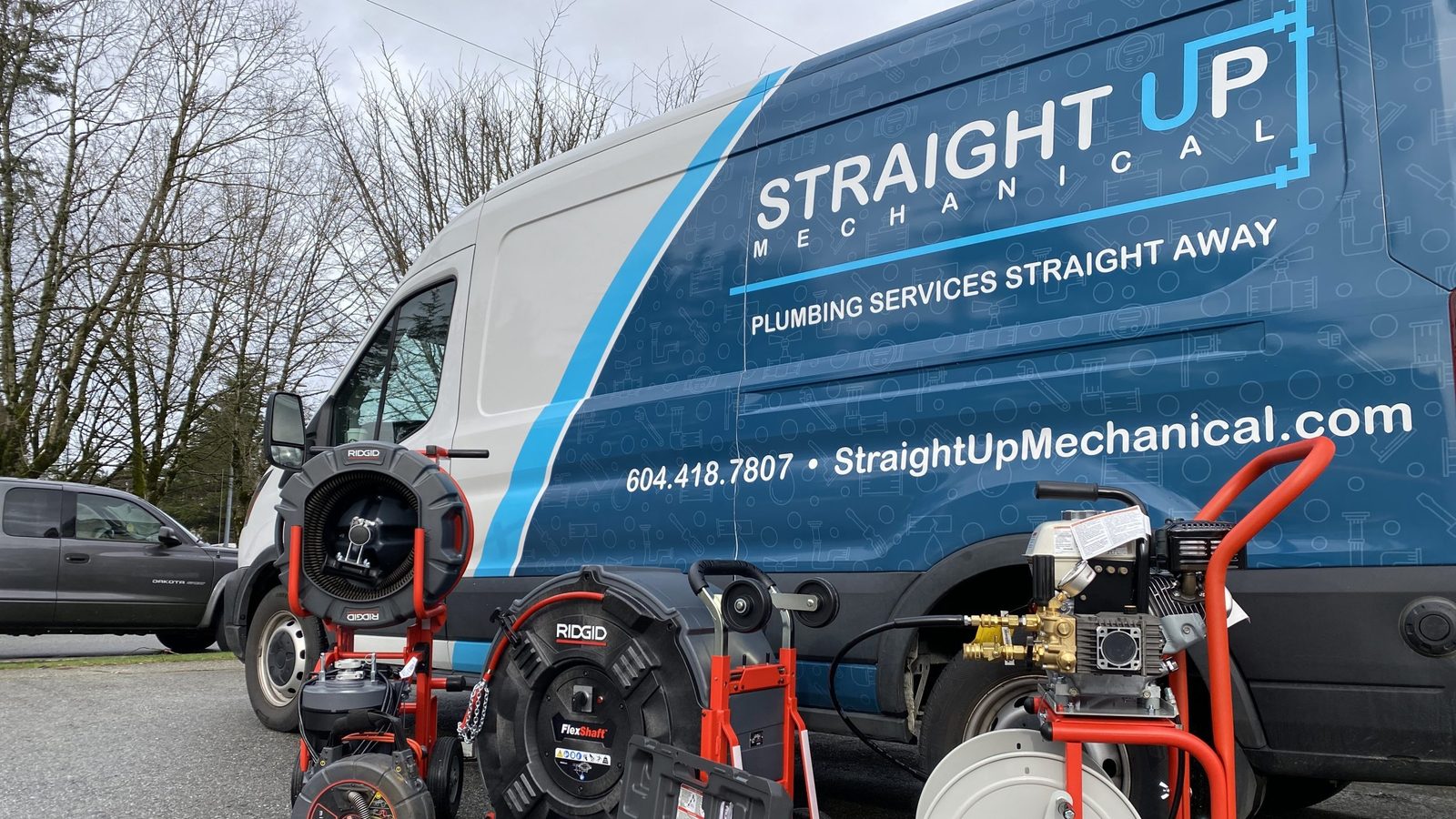 A blue and white Straight Up Mechanical van with its phone number and website on the side, parked behind drain inspection camera reels and a hydro jetter