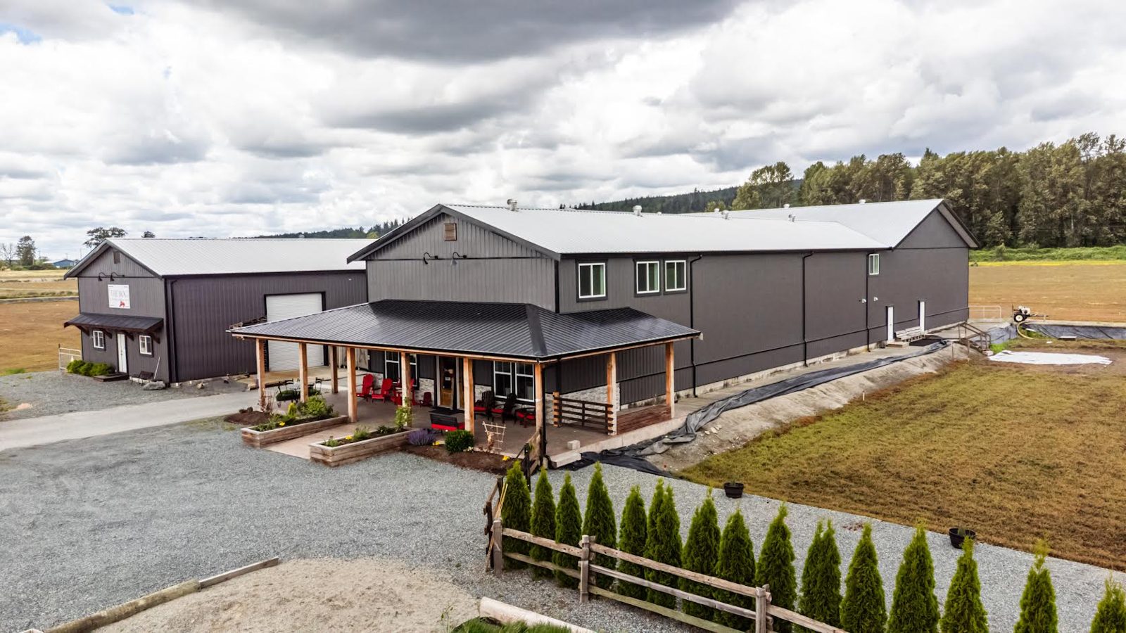 THE BOG's grey farm shop building with a covered front porch, a gravel lot and a row of young cedars, with Glen Valley fields behind