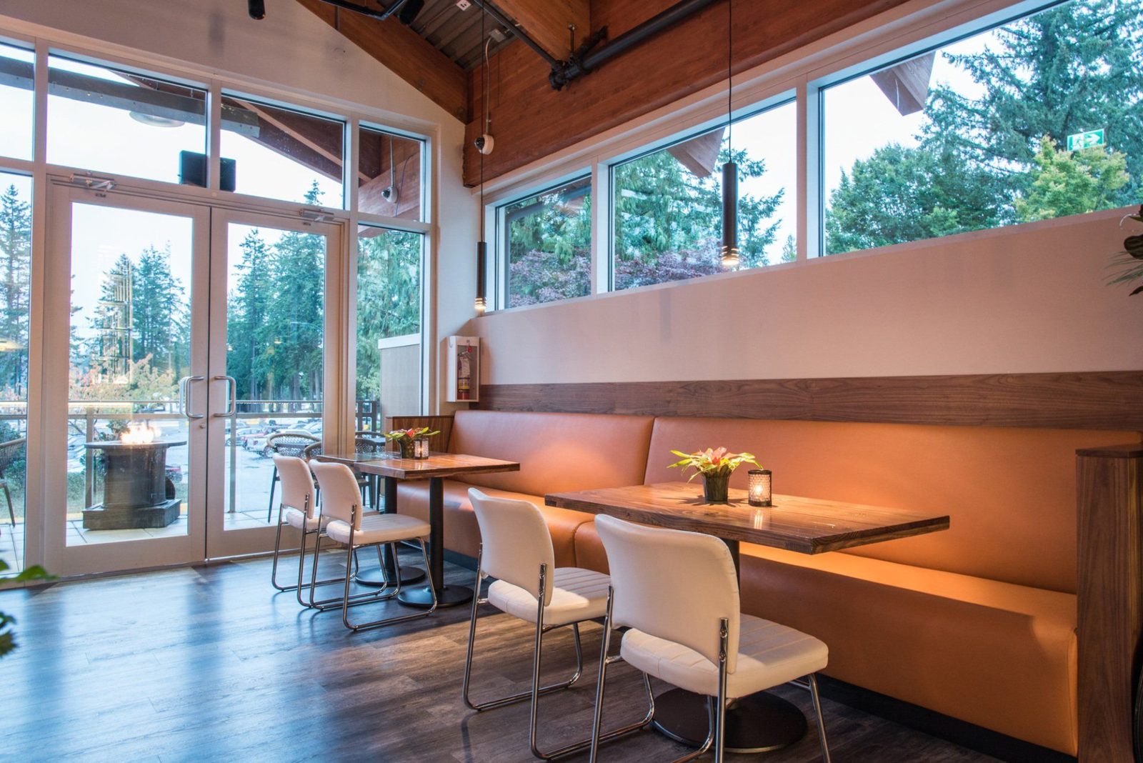 The Lodge Steakhouse dining room: a long tan leather banquette, wood tables, white chairs and tall windows looking out to evergreens and the patio fire table