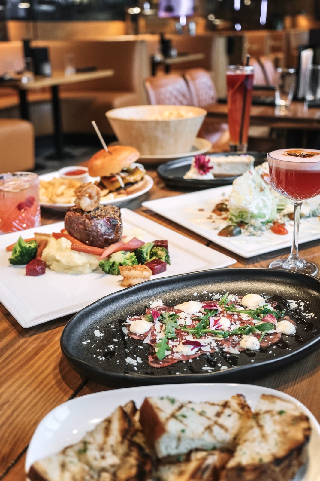A table at The Lodge set with a steak topped with a prawn, beef carpaccio, a wedge salad, a burger and fries, garlic toast and drinks