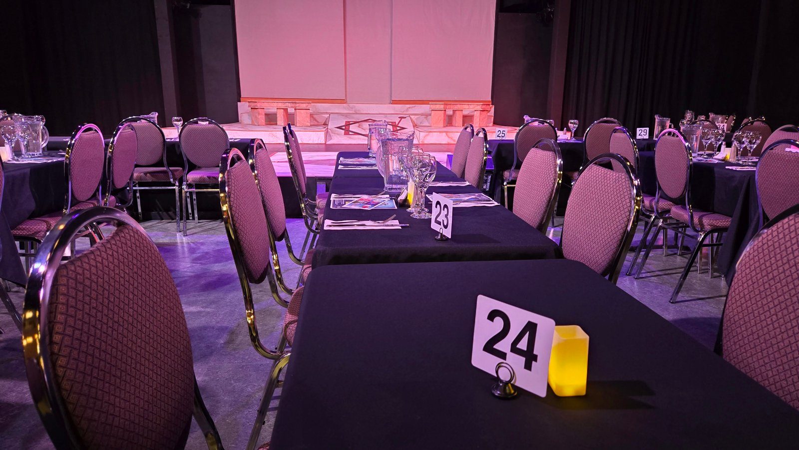 Dinner tables with black cloths and numbered cards facing the stage at Theatre in the Country, seen from table 24