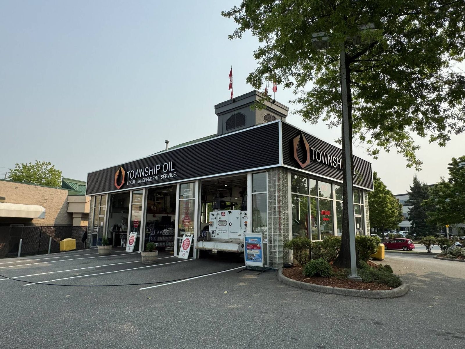 The Township Oil building on a corner lot: a black ribbed fascia with the orange drop logo and the words Township Oil, Local. Independent. Service., open service bays with a white service truck pulled in, small Canadian flags on the cupola and potted shrubs by the doors