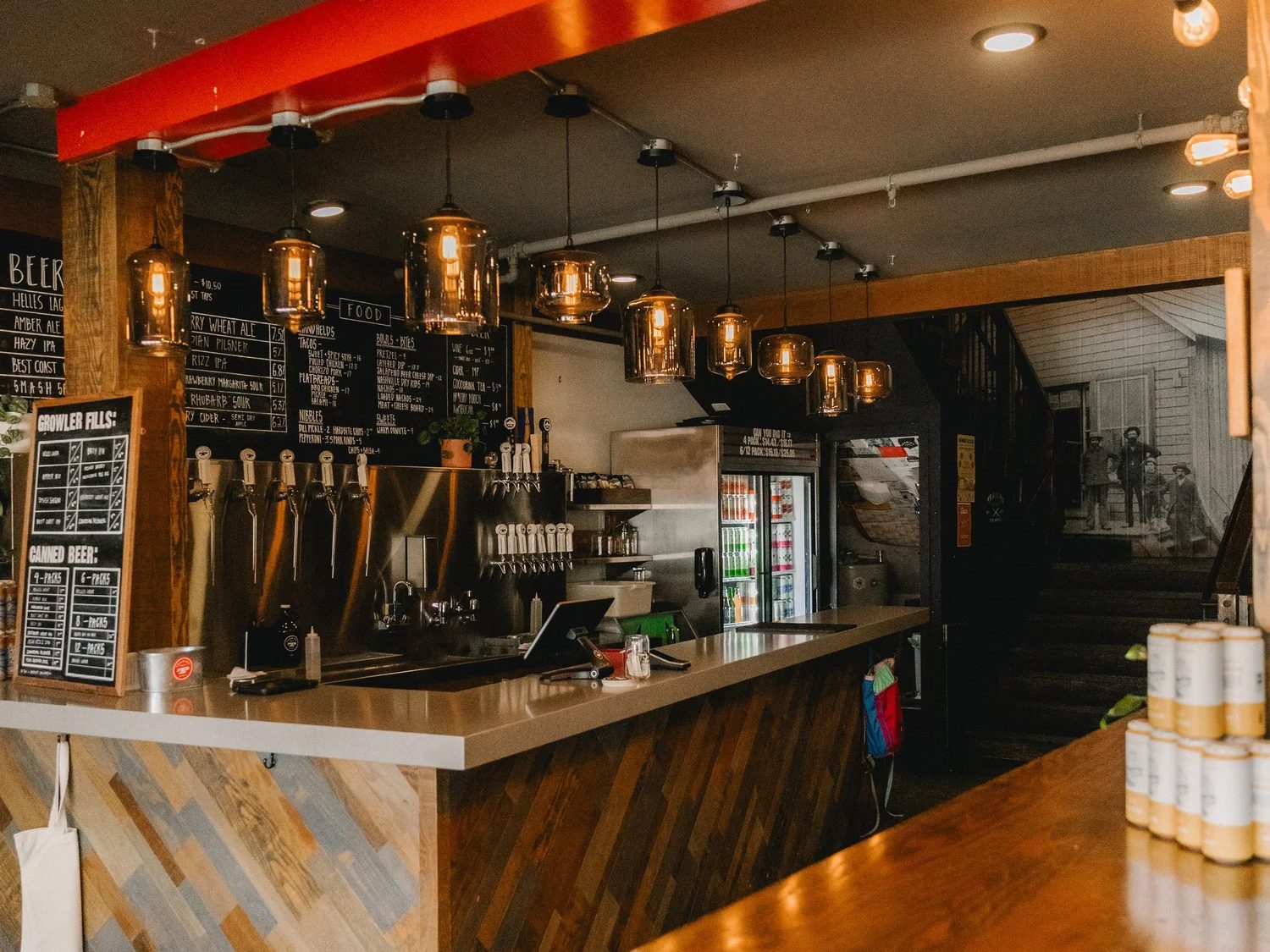The Trading Post tasting room counter: tap handles, chalkboard beer and food menus, a growler-fill board and glass pendant lights under a red beam, with stacked cans on the wood bar in the foreground