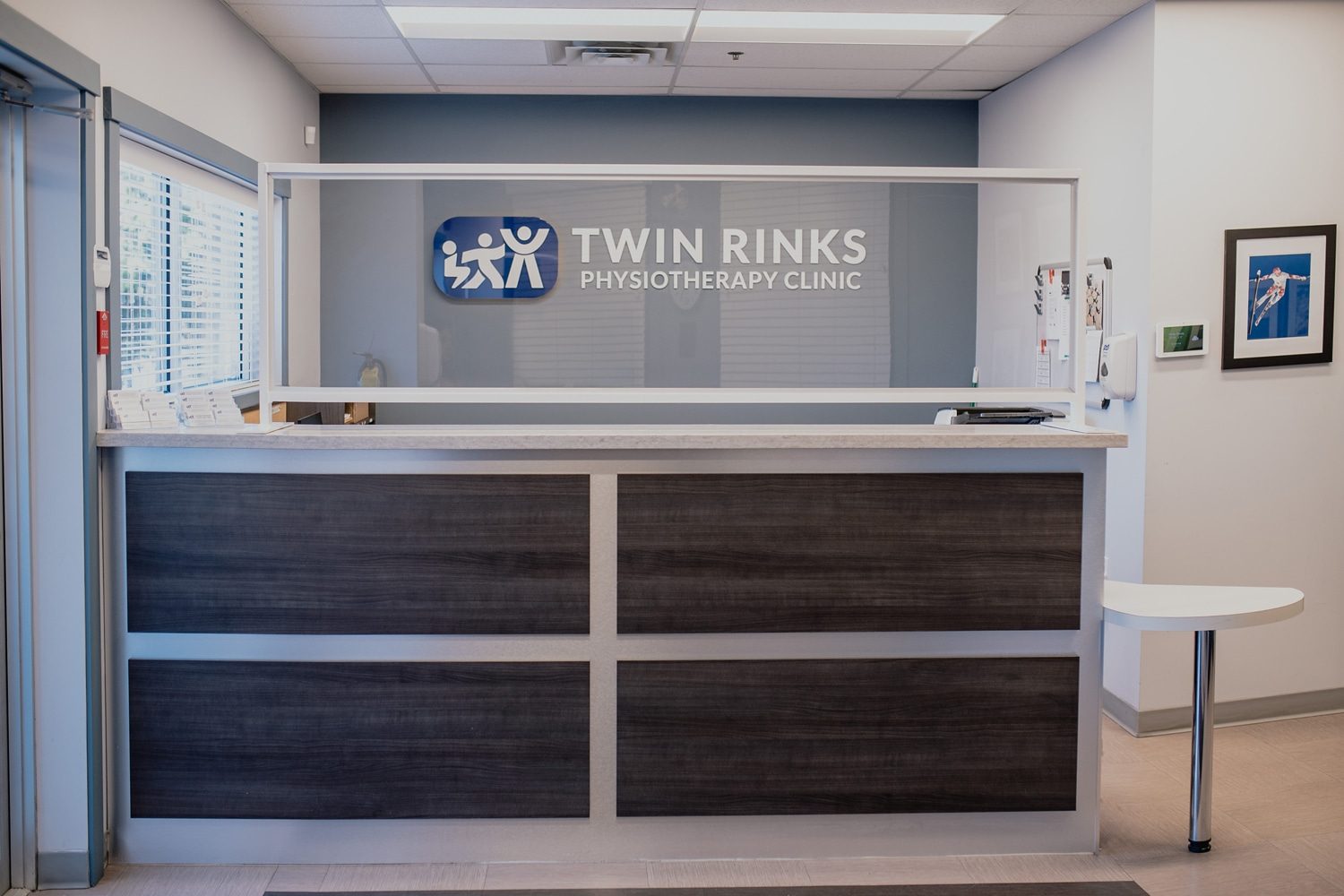 The reception desk at Twin Rinks Physiotherapy: a dark wood-grain counter under a clear screen, with the clinic's blue logo on a grey wall and a framed skiing photo to the right