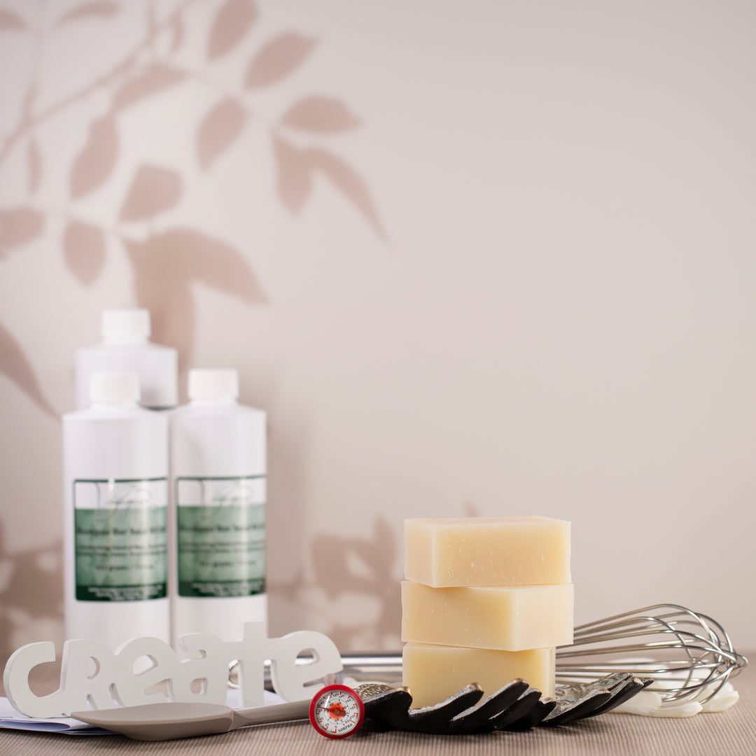 Voyageur-labelled bottles of soap-making ingredients beside a stack of plain handmade soap bars and a whisk
