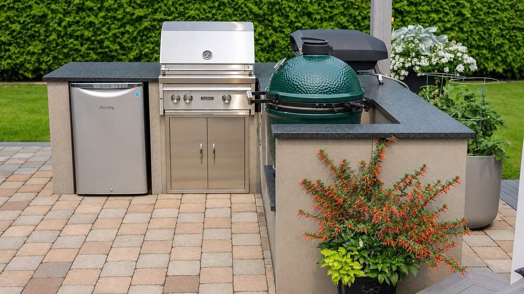 A finished outdoor kitchen on a paving-stone patio: a stainless steel built-in grill, a bar fridge and a green ceramic kamado cooker set in a stucco counter with a dark stone top, in front of a green hedge, with an orange-flowered plant in a pot beside it
