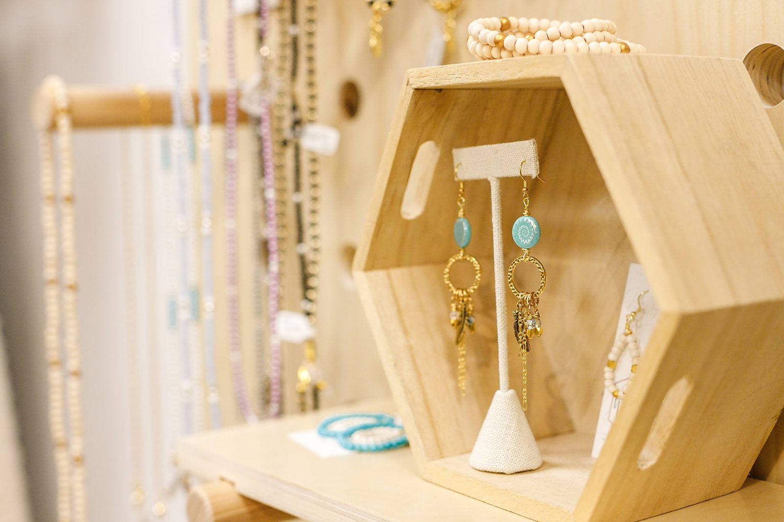Turquoise drop earrings on a small stand inside a wooden hexagon shelf, with beaded necklaces hanging behind, at Wildflowers Style & Co.