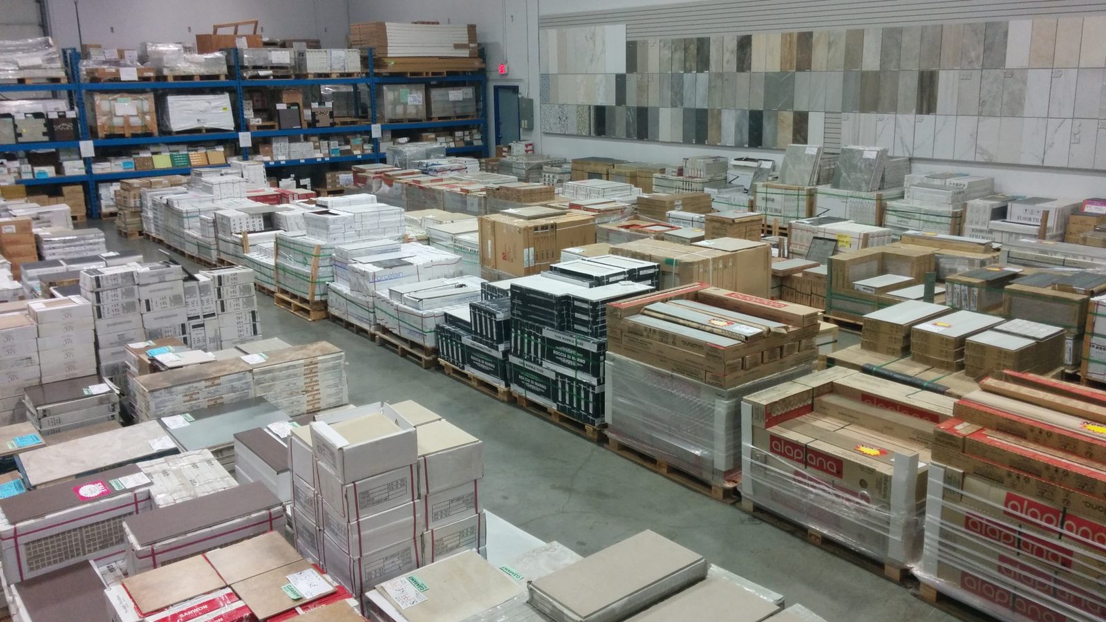 Rows of boxed tile stacked on pallets across the concrete floor of the Wilkinson Tile & Stone warehouse, with blue steel shelving and a wall of full-size tile samples at the back