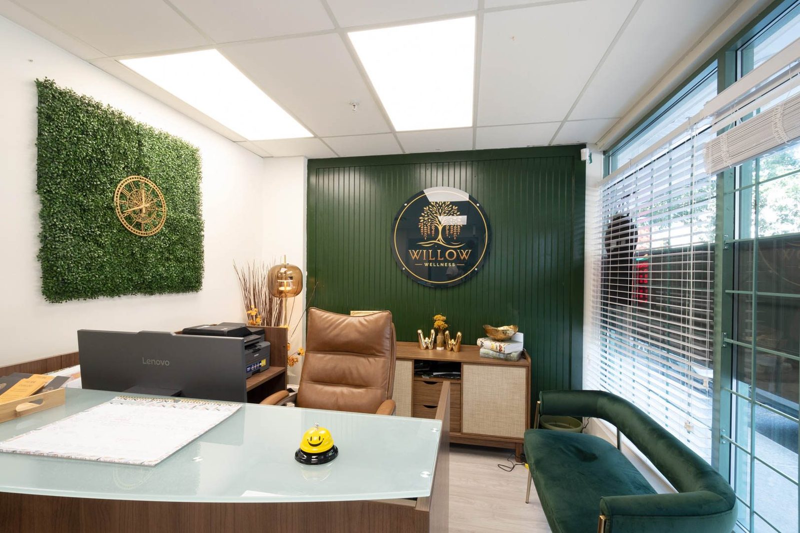 The front desk and waiting area at Willow Wellness: a green feature wall with the clinic's round willow-tree logo, a living-plant wall panel, a glass-topped desk with a small bell, and a green velvet bench by the window