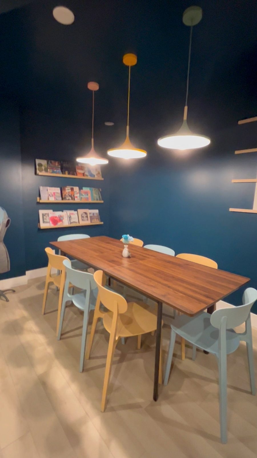 The Woolly Bulldog's workshop corner: a long wooden table with pastel chairs under pendant lights, against a deep blue wall with craft books on small shelves