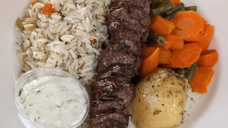 A Zythos souvlaki plate: a grilled meat skewer with rice, carrots and green beans, a roast potato and tzatziki