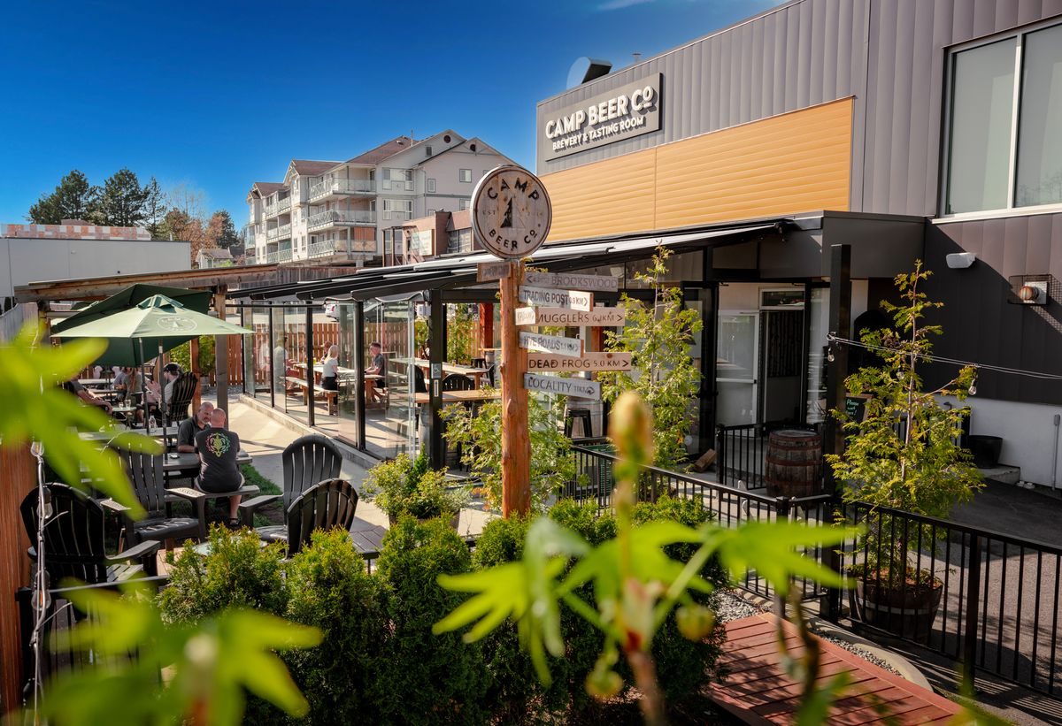Camp Beer Co.'s sunny patio with umbrellas, plants and a post clock, next to the brewery's grey building with the Camp Beer Co. sign
