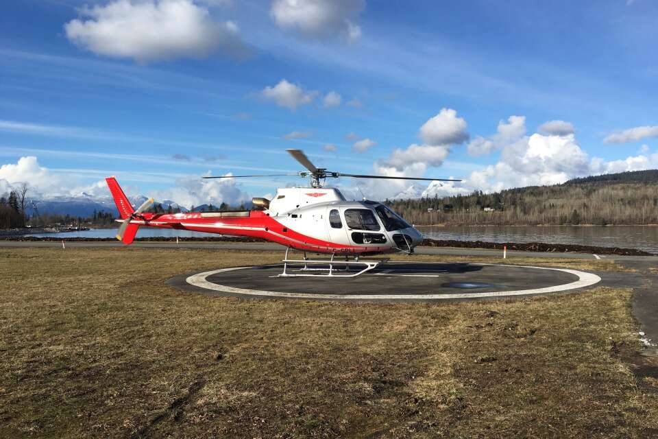 A red and white helicopter parked on a round helipad on the grass beside the Fraser River