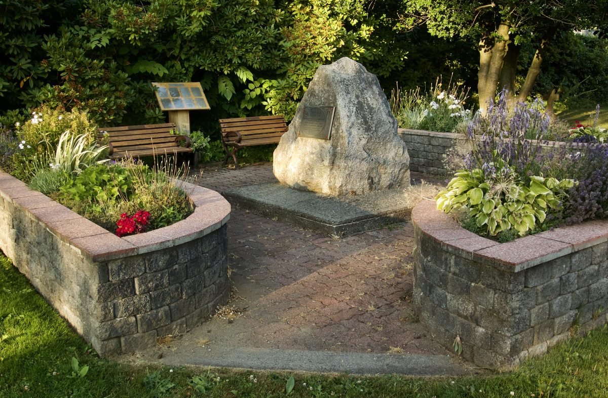 A stone memorial with benches and flower beds inside a low stone-walled garden at the Hudson's Bay Company Pioneer Cemetery in Fort Langley, where Ovid Allard is buried