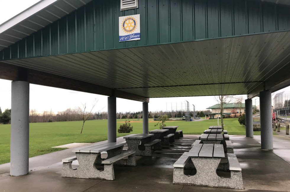 A covered picnic shelter with concrete tables at Aldergrove Athletic Park, with playing fields behind
