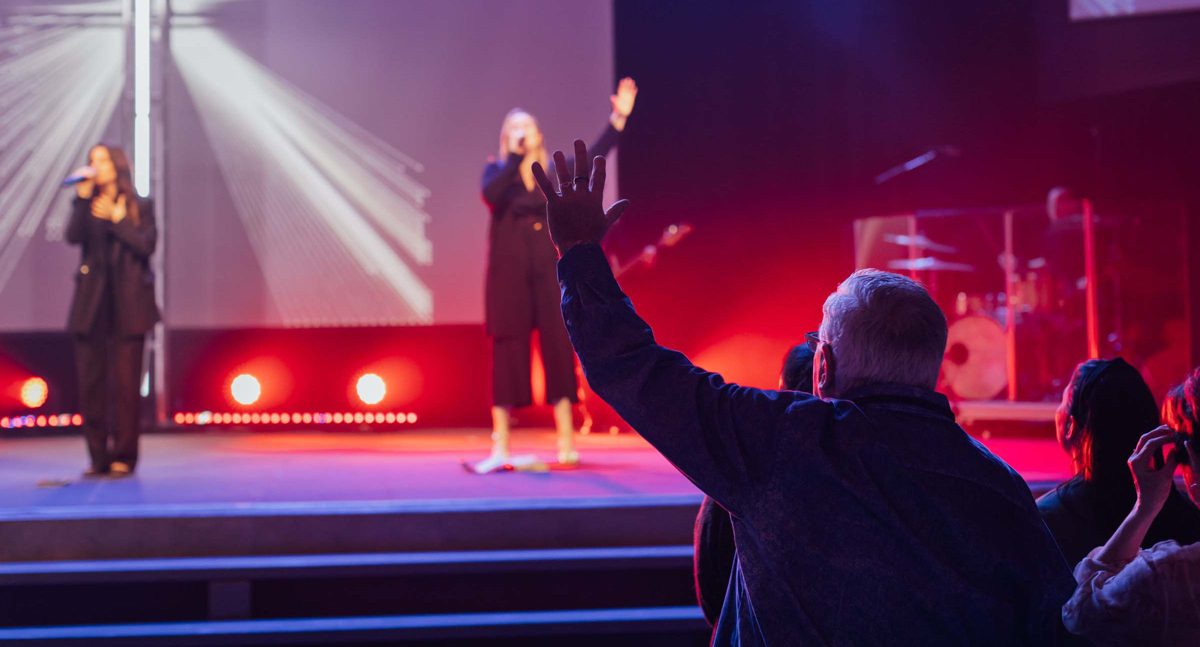 Worship leaders singing on a lit stage during a Village Church service, with people in the seats raising their hands