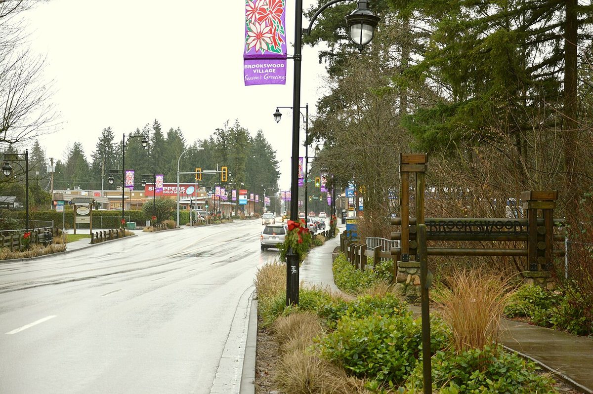 Brookswood Village on 200 Street in Langley, with decorated light poles, a log-style neighbourhood sign, a sidewalk and tall Douglas firs along the road