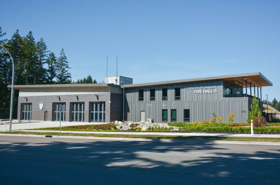 The Township of Langley's Fire Hall 5 on 27 Avenue, a two-storey grey and brick building with a wooden canopy and landscaped rocks, with tall evergreens behind