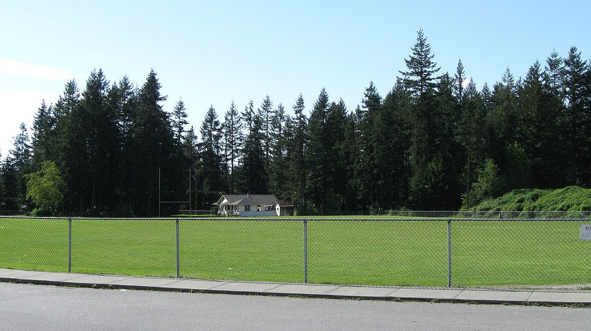 A grass field behind a Brookswood school with a house tucked among a wall of tall Douglas fir and cedar trees