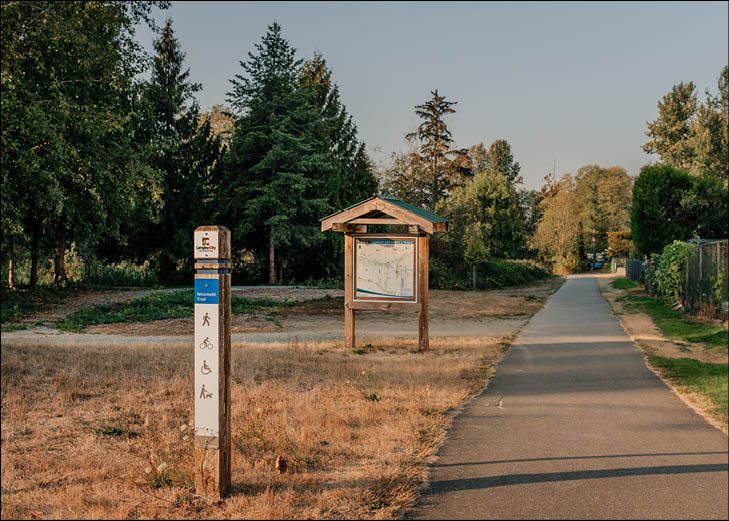 A paved Nicomekl floodplain trail in Langley City with a trail map kiosk, evergreen trees and a bench