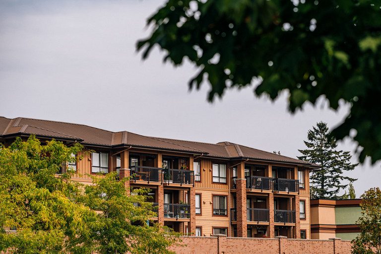 A four-storey wood-and-brick apartment building with a metal roof and balconies, seen through trees at the Village at Thunderbird in Walnut Grove