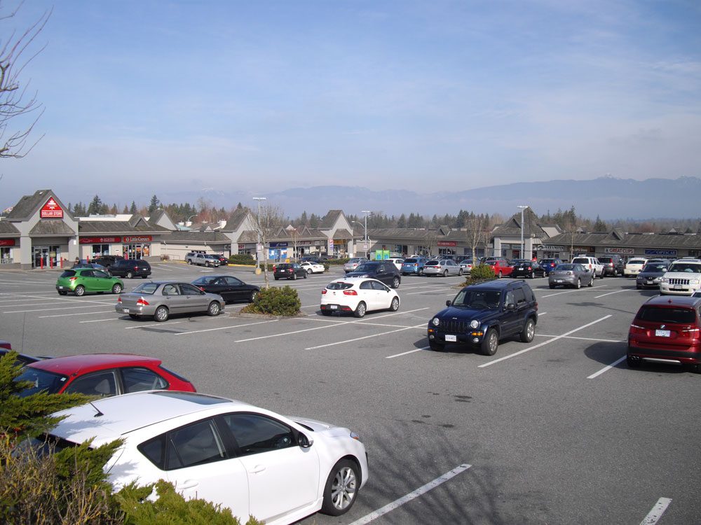 Walnut Grove Town Centre shopping plaza seen across its parking lot, with a Dollar Store and other shops under pitched roofs and mountains behind
