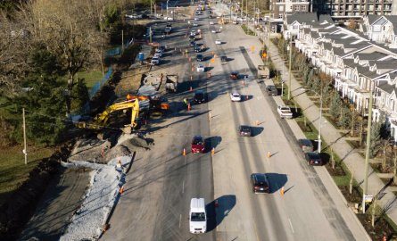 Aerial view of 208 Street widening work, with an excavator beside the road, traffic cones and a new townhouse row along the right-hand side
