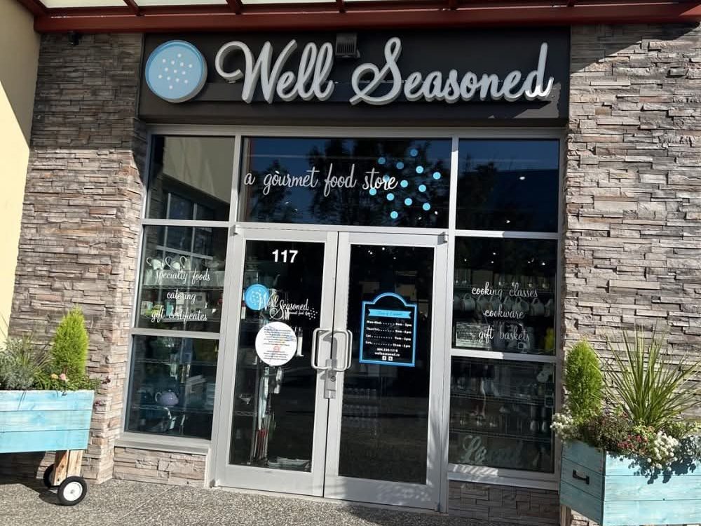 The Well Seasoned gourmet food store entrance in a 64 Avenue plaza in Willoughby, with a stone facade, glass doors and planters out front
