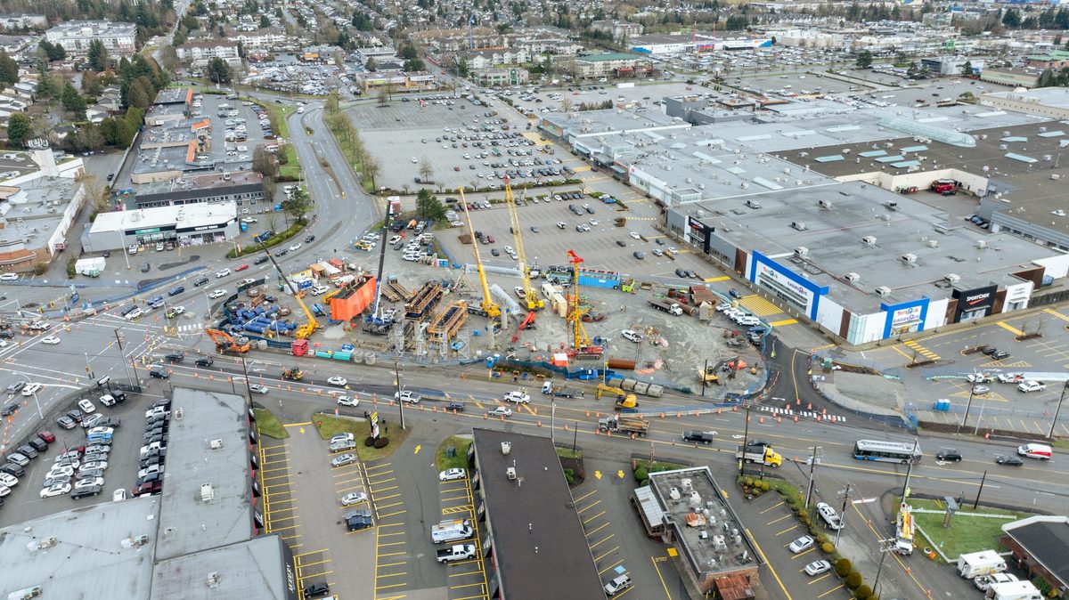 Aerial view of the Willowbrook SkyTrain station site under construction beside the parking lots of Willowbrook Shopping Centre, with cranes and traffic cones along Fraser Highway, February 2026