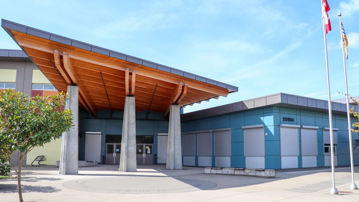 The main entrance of Yorkson Creek Middle School on 84 Avenue in Yorkson, with a sloping timber canopy over concrete columns, a blue-clad wing and flagpoles