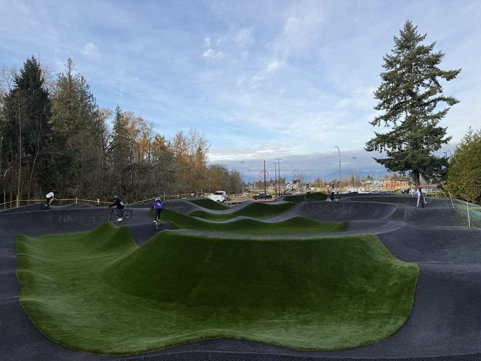 Kids on bikes and scooters riding the asphalt pump track at Yorkson Community Park, with grass-covered mounds between the lanes and tall evergreens behind