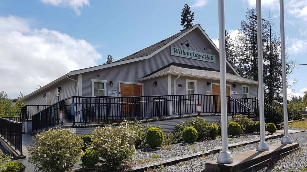 Willoughby Community Hall on 83 Avenue in Yorkson, a grey-sided hall with a wheelchair ramp, orange doors, trimmed shrubs and three flagpoles