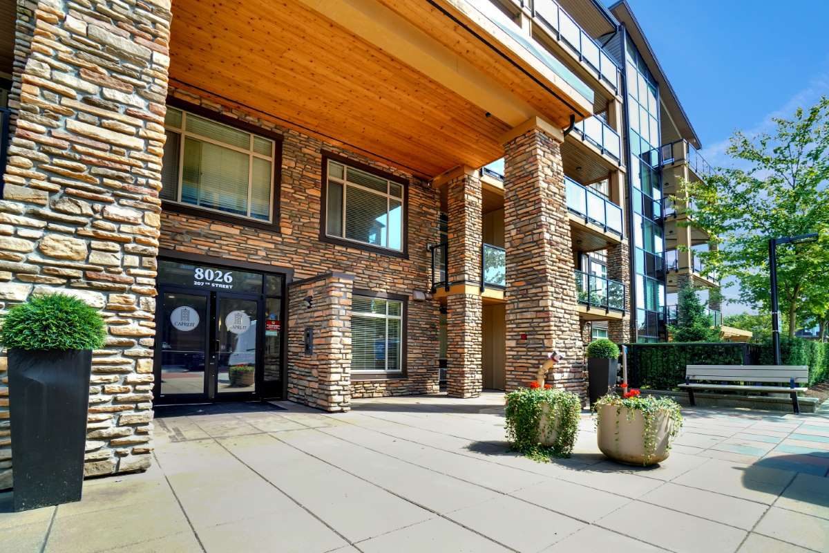 Stone-and-timber entrance of the apartment building at 8026 207 Street in Yorkson, with planters, a bench and balconies above